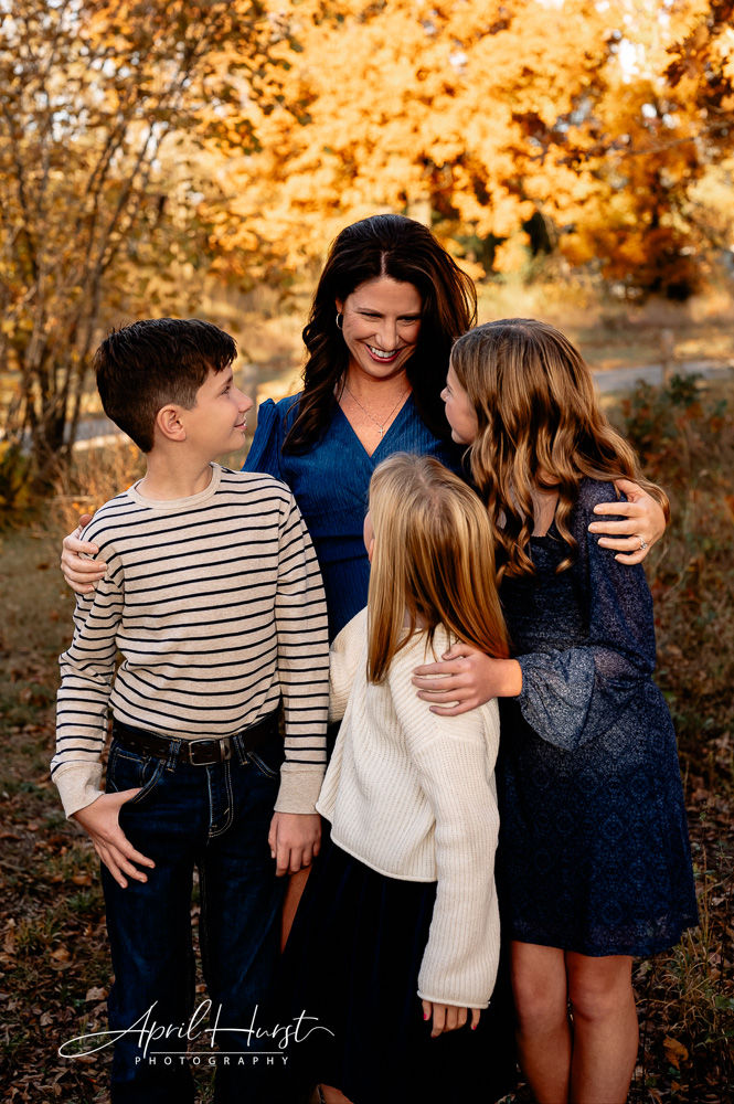 A woman embraces three children in a park with golden autumn leaves. They are smiling, wearing cozy outfits.
