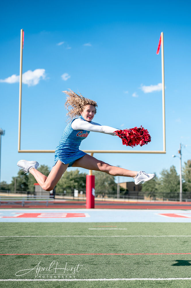 Cheerleader in blue uniform jumps with red pom-poms on a football field. Clear blue sky, goalpost, and trees in background. Energetic mood.