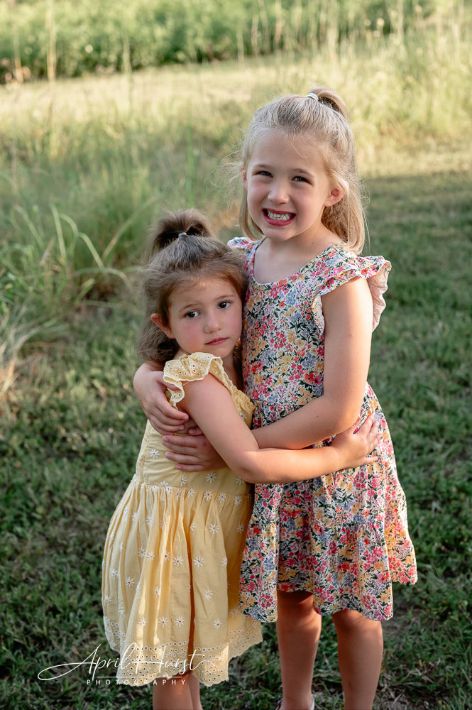 Two young girls in floral dresses hug in a grassy field. The older girl smiles, while the younger looks pensive. Warm, sunny setting.