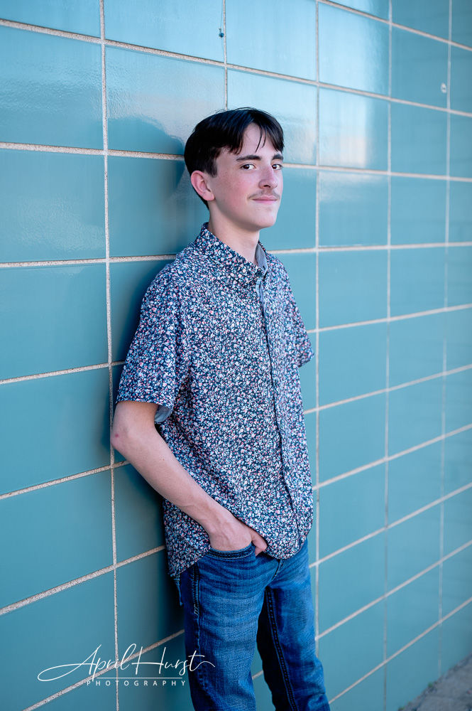 Young man in a floral shirt leans against a teal tiled wall, hands in pockets, with a confident expression.