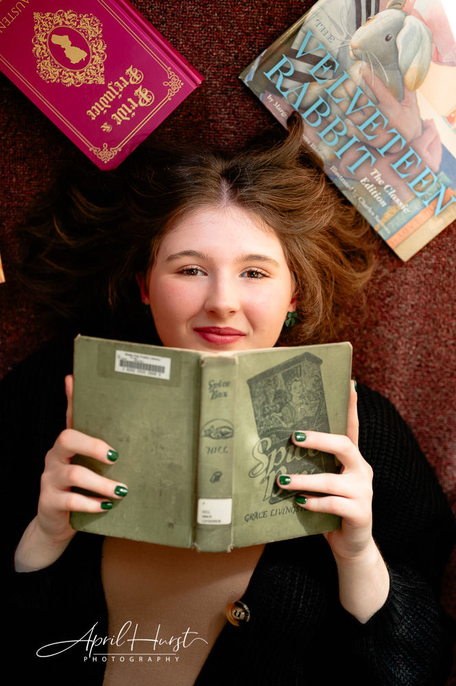 Smiling person lying on carpet, holding a green book. Surrounding books include "The Velveteen Rabbit." Cozy and relaxed mood.