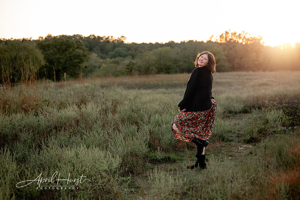 A woman in a floral skirt and black jacket smiles and twirls in a grassy field at sunset. Trees line the horizon. Mood is joyful and serene.