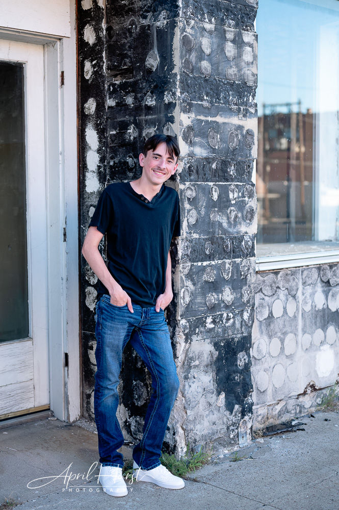 Young man smiling, leaning against a textured black-and-white wall, hands in pockets. Wears black shirt, blue jeans, white sneakers. Urban setting.