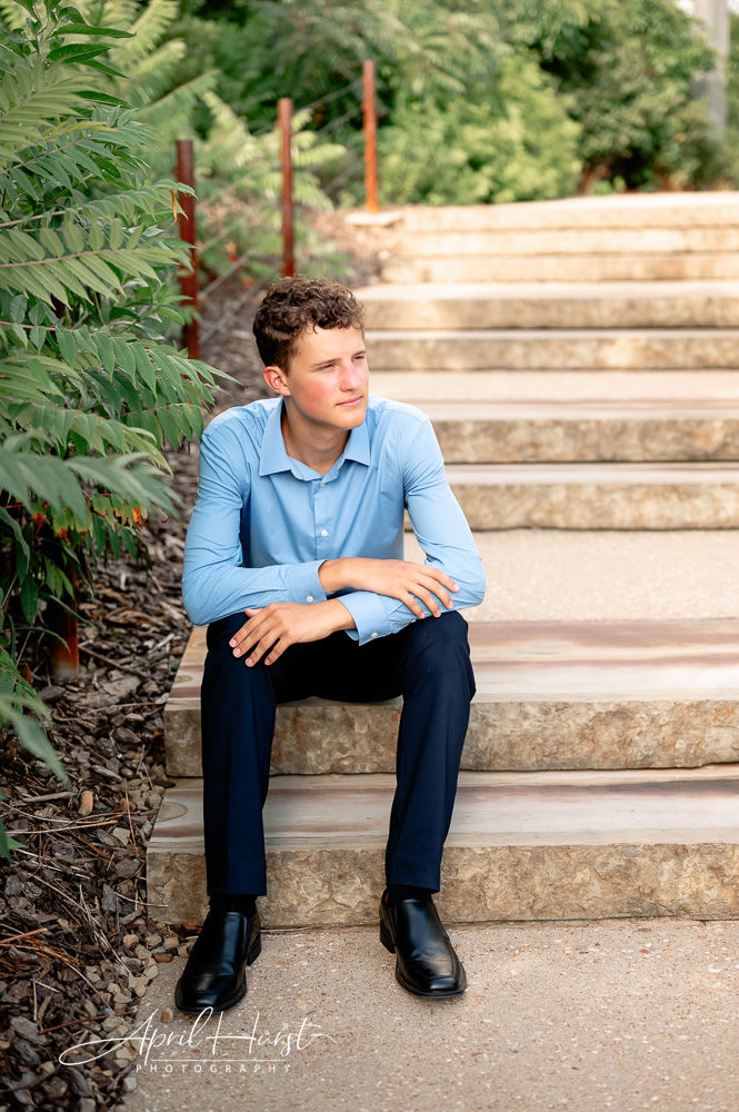 Young man in a blue shirt sits thoughtfully on stone steps, surrounded by greenery. Bright and serene outdoor setting.