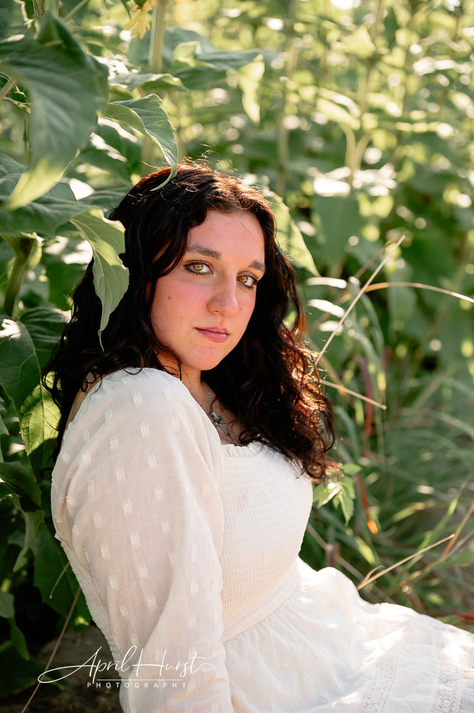 Woman in white dress sits among green leaves, looking at the camera. Sunlight highlights her face, creating a serene mood.