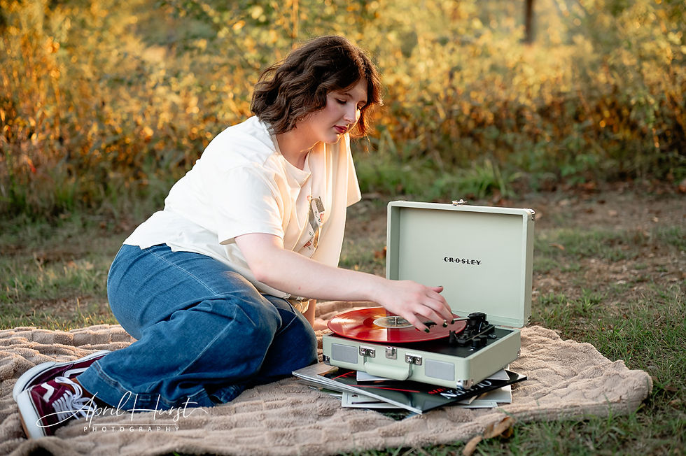 A person in a white shirt and jeans plays a red vinyl on a Crosley record player outdoors, sitting on a blanket with autumn foliage.