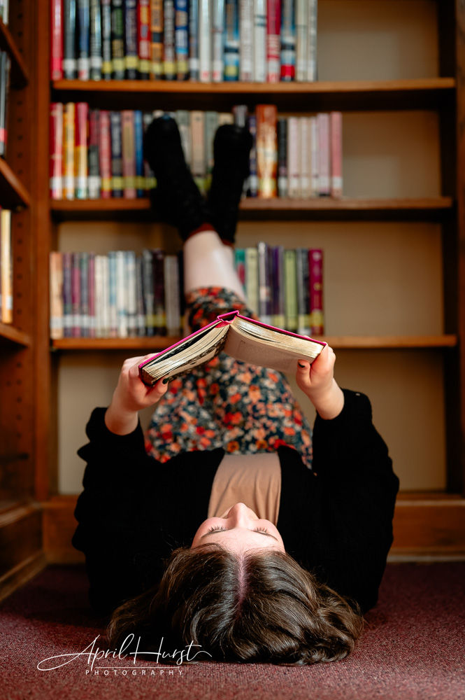 A person lies on their back in a library, legs on a bookshelf, reading a book. They wear a floral skirt, creating a relaxed mood.
