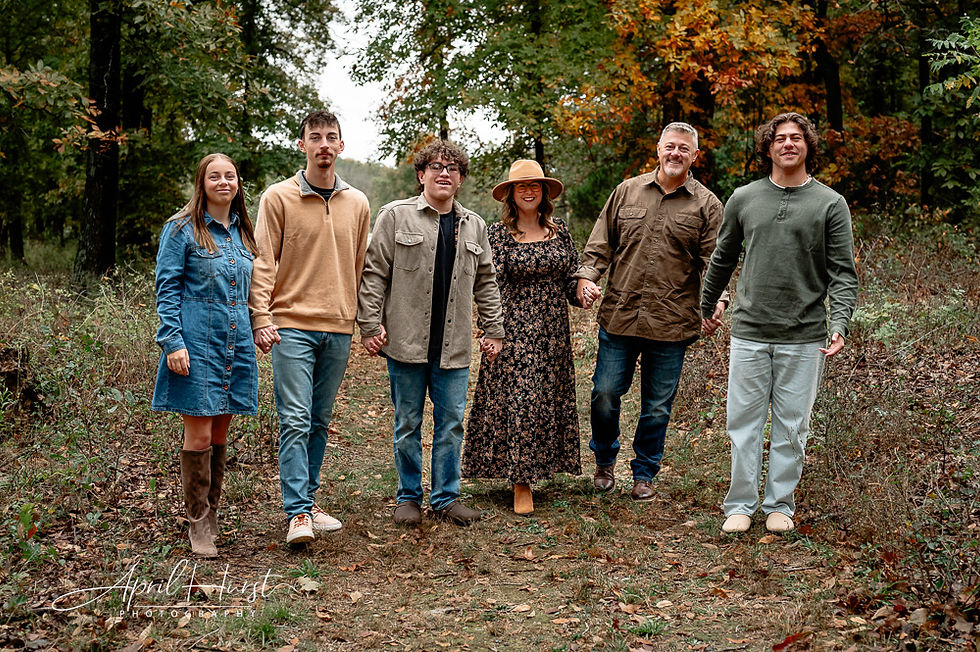Family of six holding hands, smiling in autumn forest. Casual attire with earthy tones. Lush foliage and fallen leaves in the background.