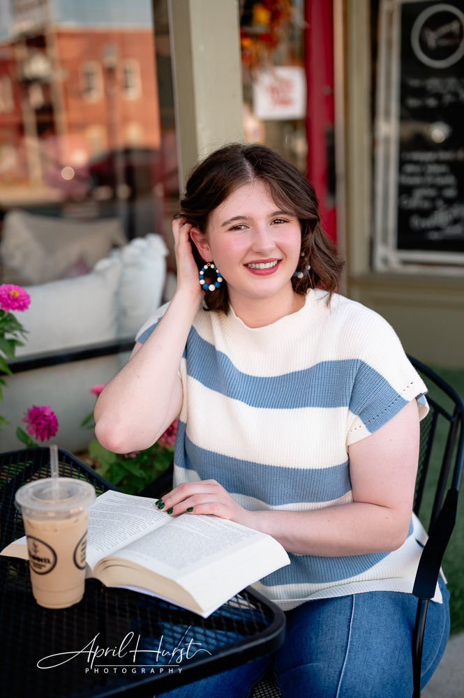 Smiling woman in striped shirt reading outdoors at a cafe table. An iced coffee and flowers are nearby. Relaxed, cheerful atmosphere.