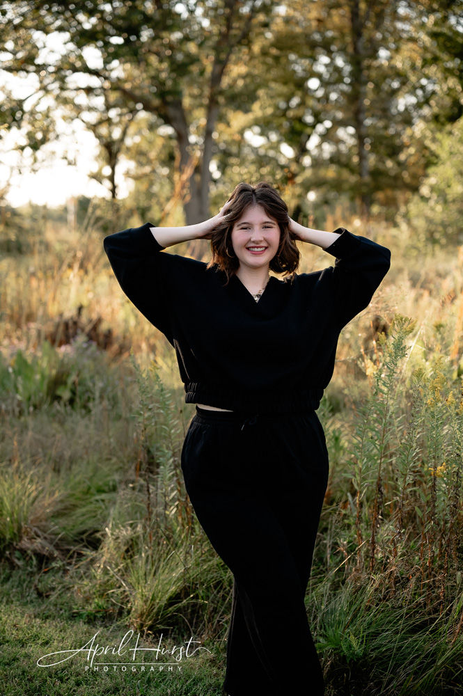 Woman in black outfit posing with hands on head, smiling amid grassy field. Sunlit trees in the background; mood is cheerful.