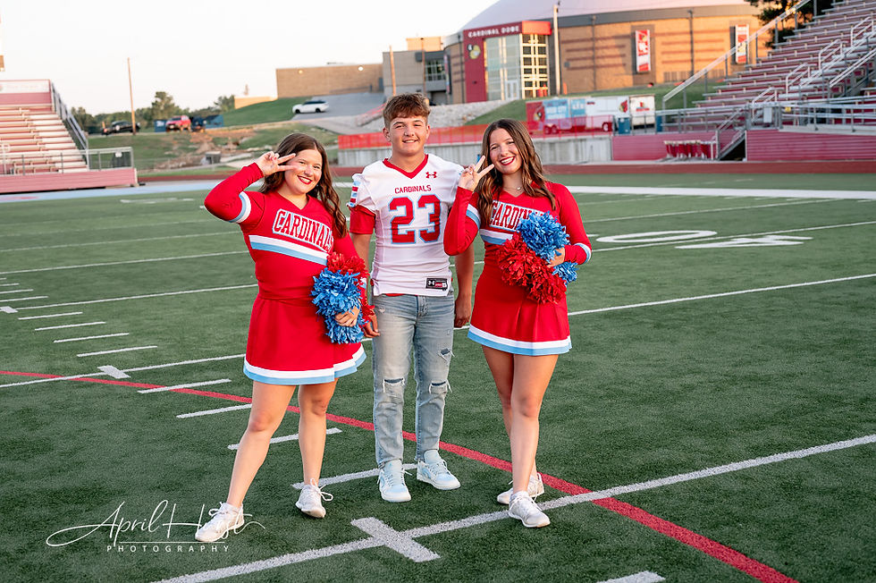 Two cheerleaders in red uniforms and a football player in jersey 23 pose on a field. Smiling, colorful pom-poms, stadium backdrop.