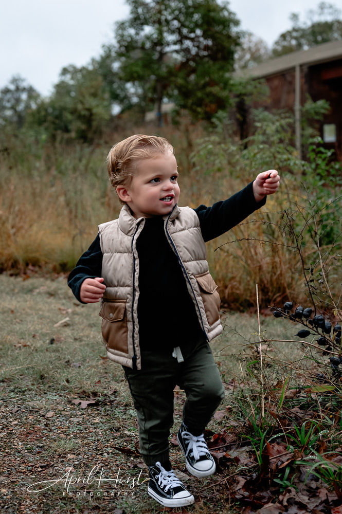 Young child in a beige vest and black shirt walks on a gravel path, smiling and reaching out. Background features greenery and a rustic building.