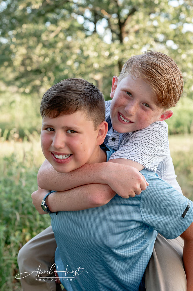 Two boys smiling while giving a piggyback ride in a sunny park. One wears blue, the other striped. Lush greenery in the background.
