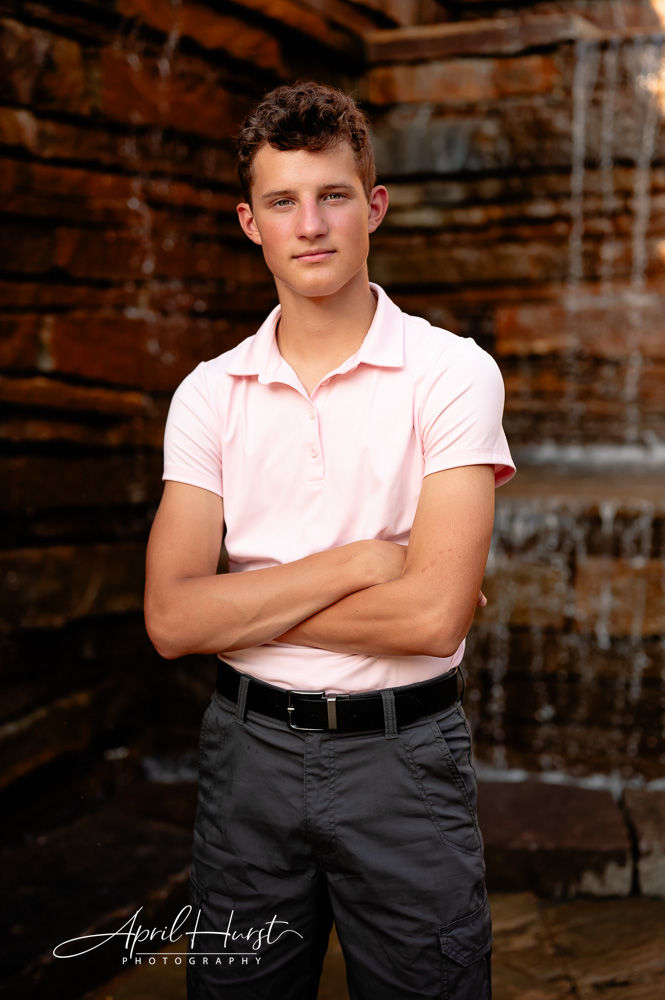 Teen in pink shirt stands with arms crossed, expression neutral. Background of rustic stone wall with waterfall.