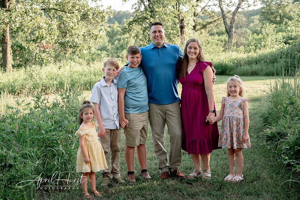 Family of six smiling, standing in a lush green field. Parents in blue and maroon, kids in pastel clothes. Bright, sunny day atmosphere.