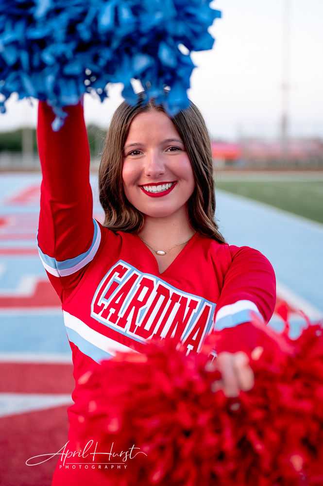 Cheerleader in red uniform with "Cardinals" text, smiling and holding red and blue pom-poms on a sports field.