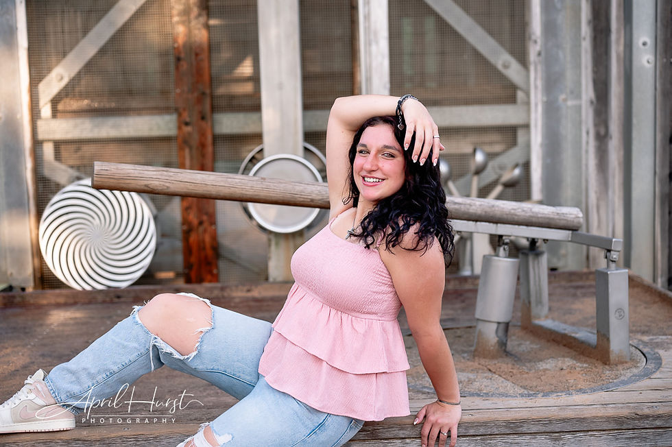 A woman in a pink top and ripped jeans poses confidently on wooden steps. Gray metallic background with artistic patterns visible.