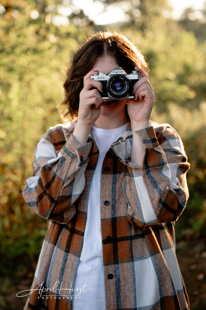 Person in plaid shirt holds a Canon camera outdoors, with blurred greenery in the background. Warm lighting creates a cozy atmosphere.