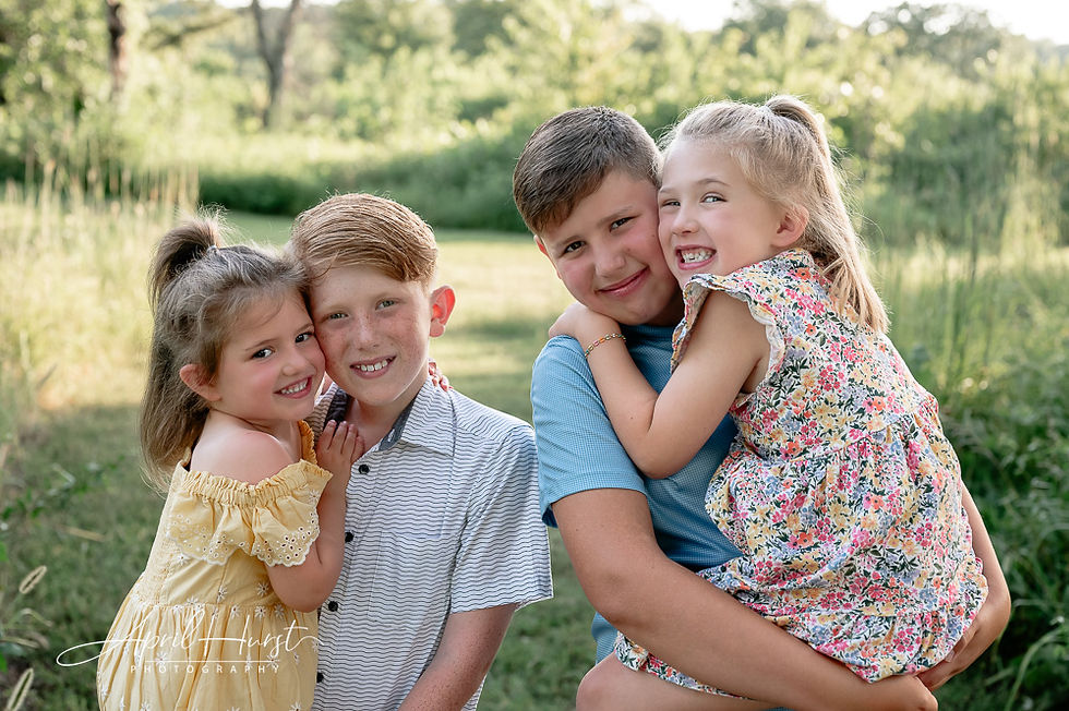 Four smiling children, two girls in floral dresses and two boys in striped shirts, stand and hug in a sunny, grassy field.