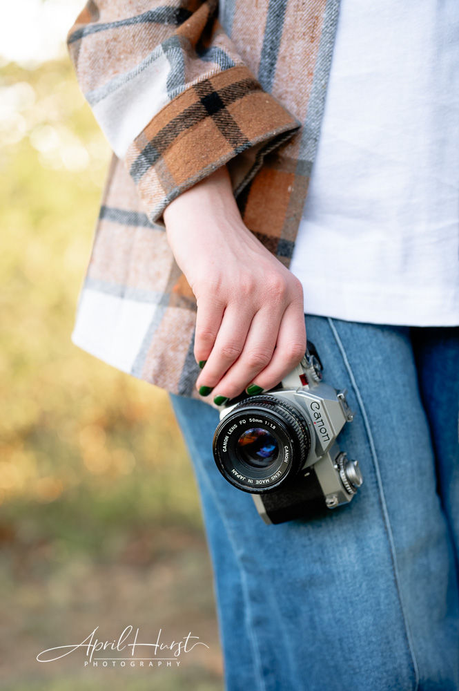 A person in a plaid shirt holding a Canon film camera stands in a sunny outdoor setting. Jeans and a white shirt are visible. Mood is calm.