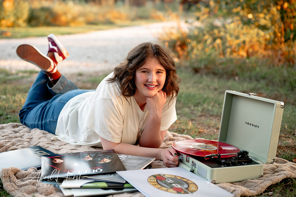 A smiling person lies on grass with a Crosley record player and albums. They're wearing jeans and sneakers. The setting is sunny and relaxed.