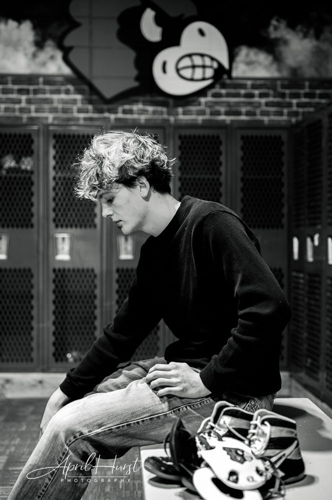Young man in a locker room sits pensively on a bench. Black and white photo with lockers and cartoon mural in the background. Football gear nearby.
