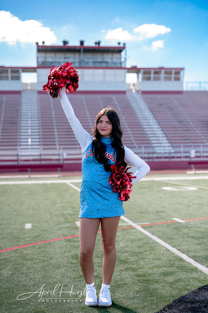 Cheerleader in blue uniform with red pom-poms raises arm on a football field. "Cardinals" text on uniform. Stadium seats in background.