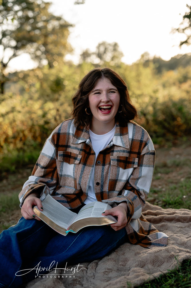 Smiling person with open book, wearing a plaid shirt, sitting on a blanket outdoors. Background features trees and sunlight.