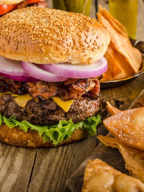 Juicy cheeseburger with bacon, lettuce, and onion on a sesame bun, surrounded by crispy chips. Rustic wood table, fresh tomatoes nearby.