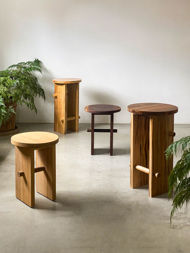 Banquetas Trini em madeira maciça clara e escura, ao lado de planta e estante modular