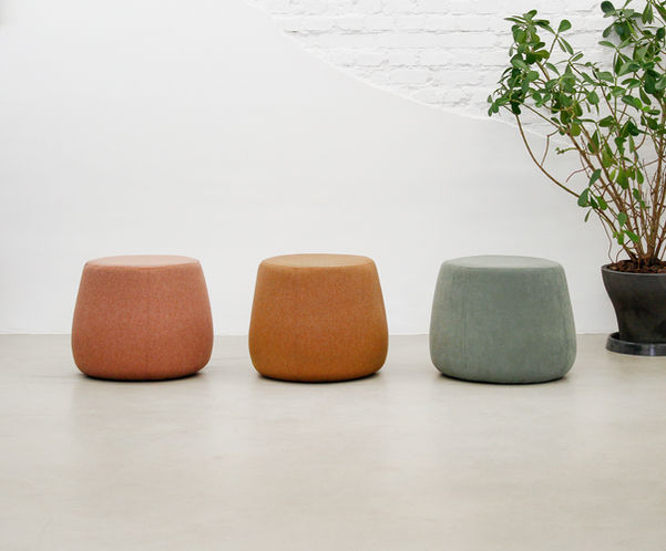 Três Puffs Cícero enfileirados em tecido rosa-terracota, mostarda e camurça verde, diante de parede de tijolo branco