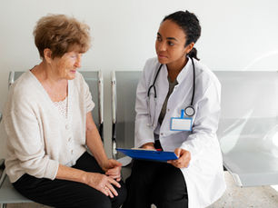 Senhora branca de cabelo dourado sentada numa cadeira sendo atendida por uma médica negra de jaleco e prancheta azul