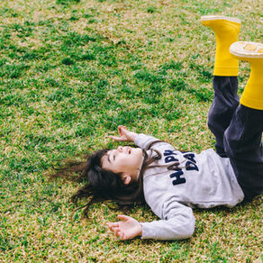 Criança deitada na grama com botas amarelas, calça preta e moletom cinza.