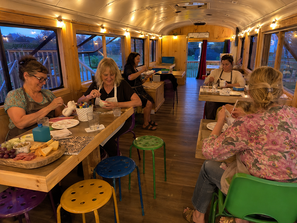 Women painting ceramic figurines at tables in a cozy wooden bus studio at dusk, with colorful stools and a Sortie de secours sign.