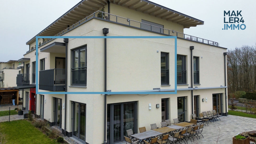 Außenansicht des Gebäudes aus einem anderen Winkel. Die blau markierte Wohnung im 1. Obergeschoss verfügt über einen Balkon mit Metallgeländer. Im Hintergrund ist eine weitere Wohnanlage zu sehen.