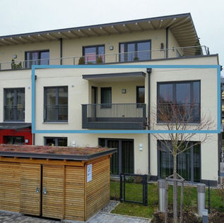 Moderne, mehrgeschossige Wohnanlage mit Balkonen und Flachdach, im Vordergrund ein Holzschuppen und gepflegter Gemeinschaftsbereich mit Sitzmöglichkeiten.