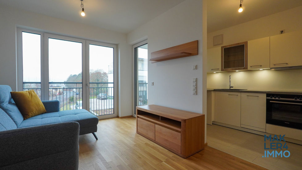 Heller Wohnbereich mit bodentiefen Fenstern und Zugang zum Balkon. Die Einrichtung umfasst eine blaue Couch mit gelbem Kissen, einen niedrigen TV-Schrank und eine schlichte Holzvitrine.