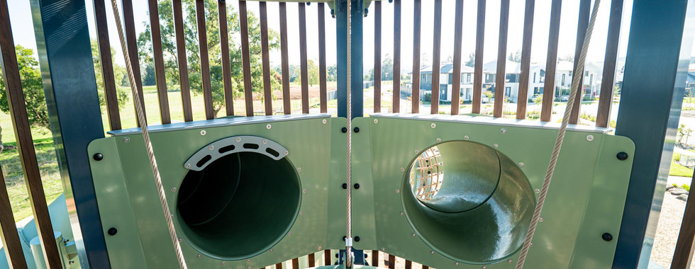 A multi-level custom timber look playground featuring two large towers linked by a suspended rope mesh tunnel, multiple tube slides, an internal rope climbing net and an elevated bridge.