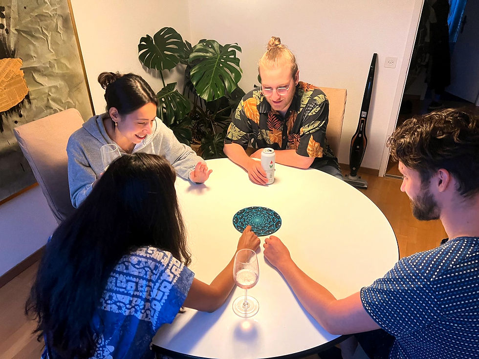 Four people smiling around a round table with drinks, one holding a can, near green plants and a vacuum. Cozy indoor setting.