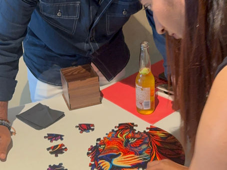 Two people assemble a colorful lion puzzle on a table. A bottle with a lime slice and a wooden box are nearby, creating a focused atmosphere.