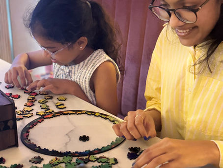 Two people assembling a colorful puzzle on a white table. One wears glasses and a yellow striped shirt. They appear focused and engaged.