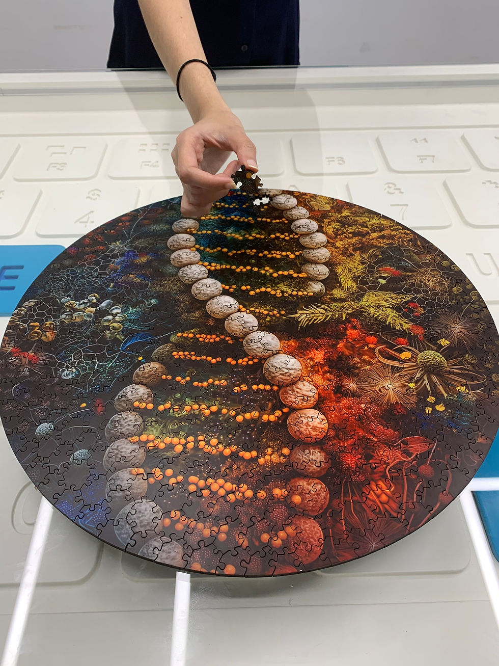 A hand places a puzzle piece on a round, colorful DNA-themed puzzle on a white table, with visible letters and numbers in the background.