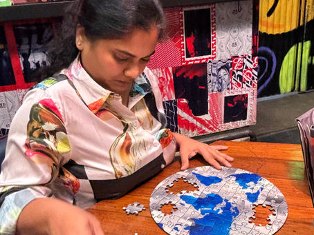 Woman in a patterned shirt assembles a round jigsaw puzzle at a wooden table in a colorful café with QR code signs.