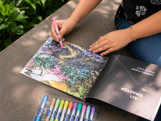 Person coloring a detailed book outdoors with a pink marker. Nearby, a set of colorful markers on a stone surface. Lush greenery in the background.
