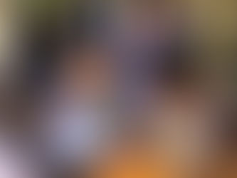 Four people engage with a wooden puzzle at a table in a warm-lit room. The background shows others socializing near green wall panels.