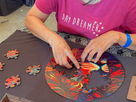 Person in a "Day Dreamer" shirt works on a colorful, circular puzzle at a table. A wooden box and extra pieces are nearby.