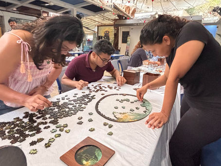 Three people are focused on assembling a circular puzzle on a table in a lively café setting, surrounded by other patrons.