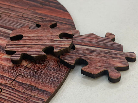 Two wood-patterned jigsaw pieces rest on a partially completed circular puzzle with matching design. Background is a neutral surface.