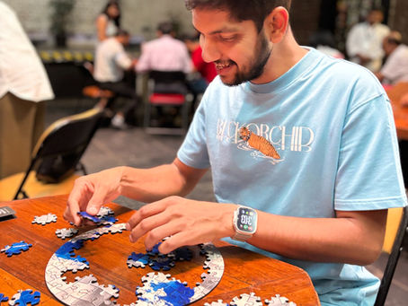 A man in a blue shirt assembles a blue and white puzzle on a wooden table in a casual indoor setting. A logo "COGZART" is visible on the wall.