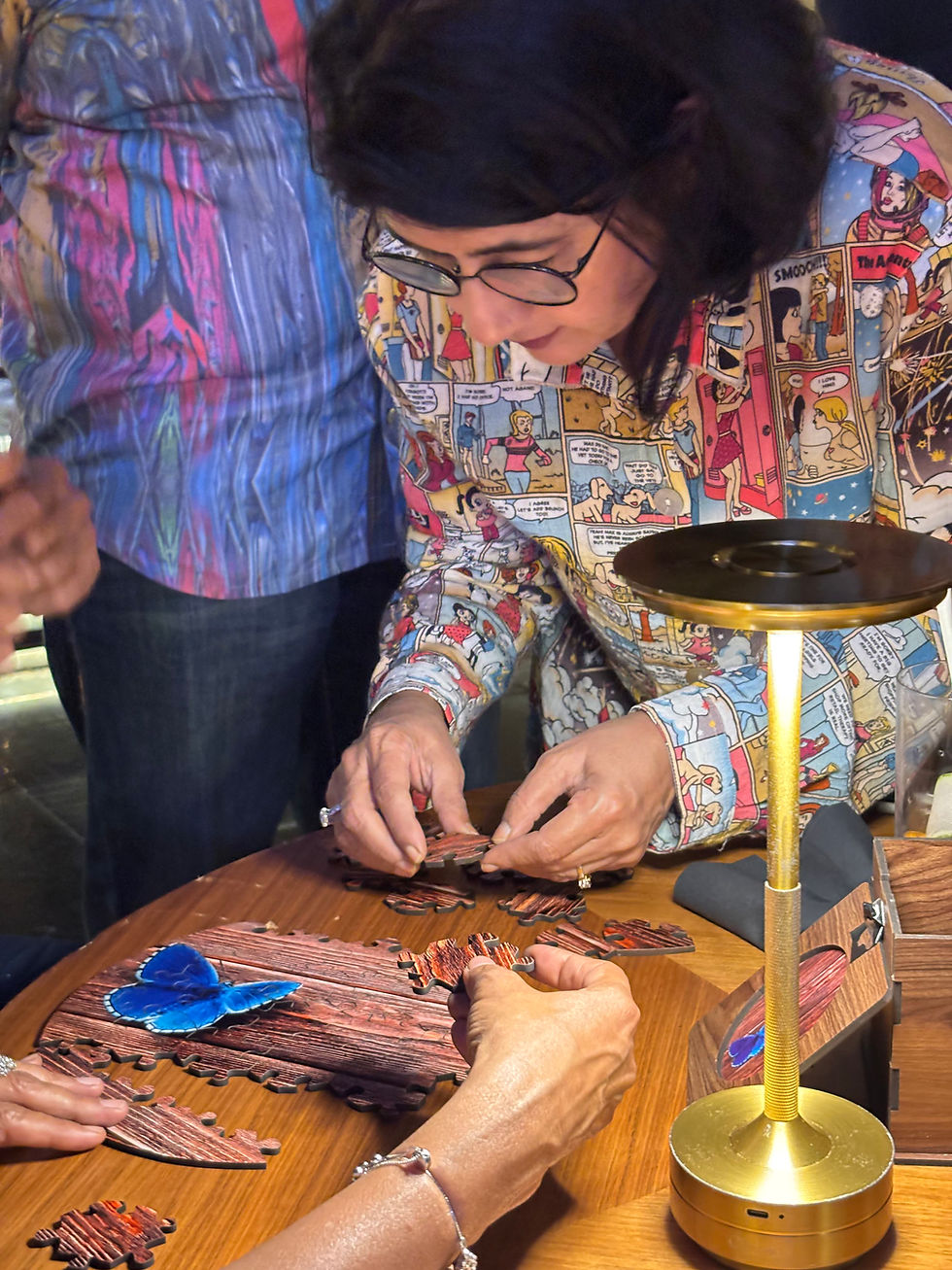 A person wearing a comic-patterned shirt assembles a puzzle with others. A blue butterfly piece is set on a wooden table under a lamp.